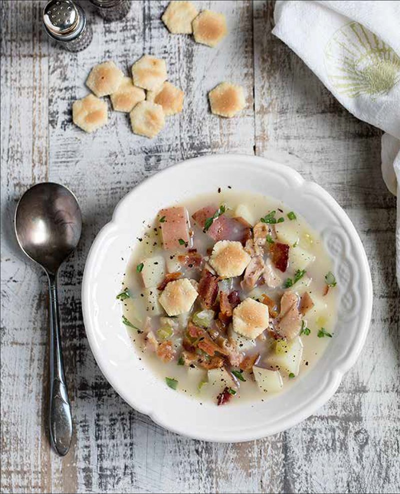 Rhode Island Clam Chowder from the Electric Pressure Cooker Cookbook - featuring overhead shot of a pale white broth with diced potatoes, bacon, parsley, cracked pepper, and oyster crackers on top