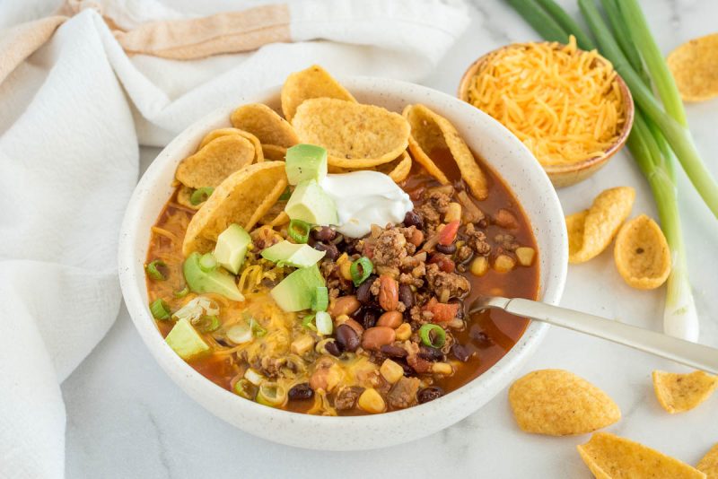 Overhead picture of Instant Pot taco soup topped with sour cream, cheese, avocado, and chips with shredded cheese and green onion in the background.