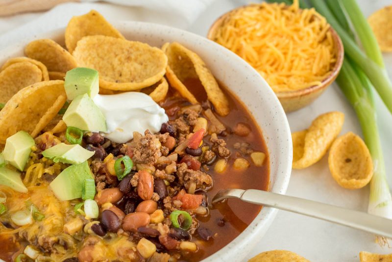 A close up picture of a bowl of Instant Pot taco soup topped with avocado, cheese, sour cream, chips, and green onion, with shredded cheese and green onion in the background.