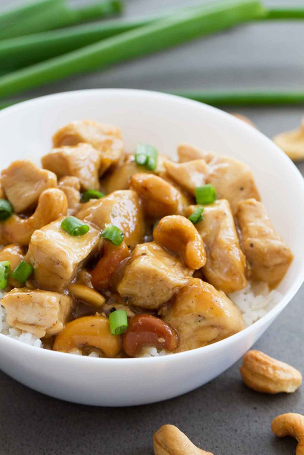 45 degree shot of Instant Pot Cashew Chicken from Taste and Tell's review of the Electric Pressure Cooker Cookbook - featuring a white bowl filled with white rice, bite-sized chicken and cashews coated in a light sauce and garnished with green onions