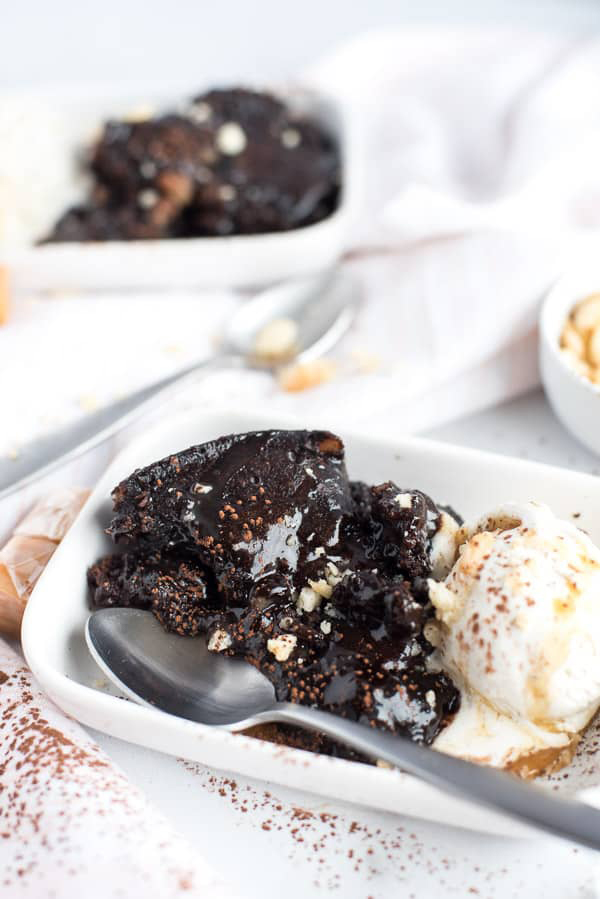 45 degree shot of Instant Pot Dark Chocolate Caramel Brownie Pudding from Tidbits, included in the Instantly Sweet Dessert Cookbook - featuring a white dish with a spoon buried in gooey dark chocolate pudding with ice cream topped with caramel sauce
