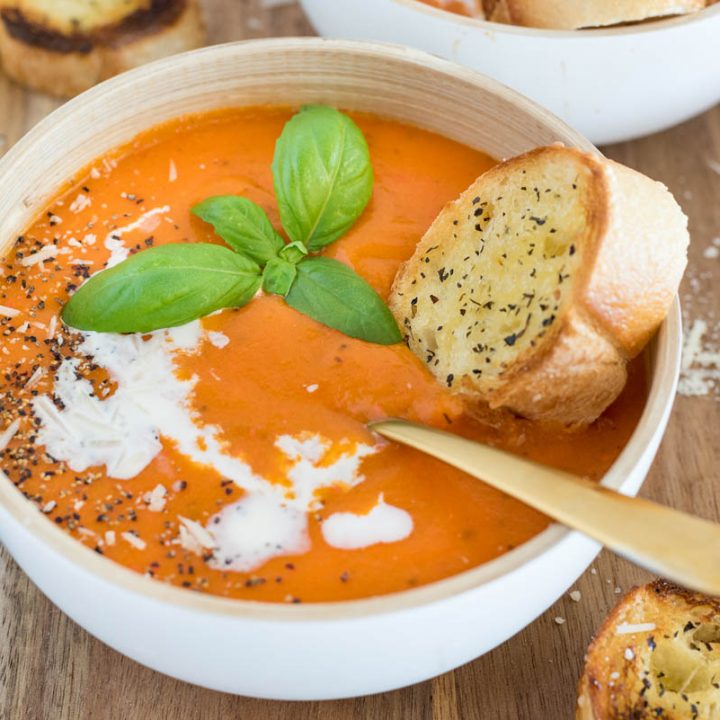 a 45 degree shot of a white bowl against a wooden background. The bowl is filled with orange tomato soup, garnished with basil, salt and pepper, and heavy cream, with a silver spoon and a baguette on the side.