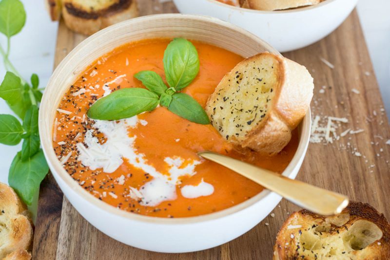 a 45 degree shot of a white bowl against a wooden background. The bowl is filled with orange tomato soup, garnished with basil, salt and pepper, and heavy cream, with a silver spoon and a baguette on the side.