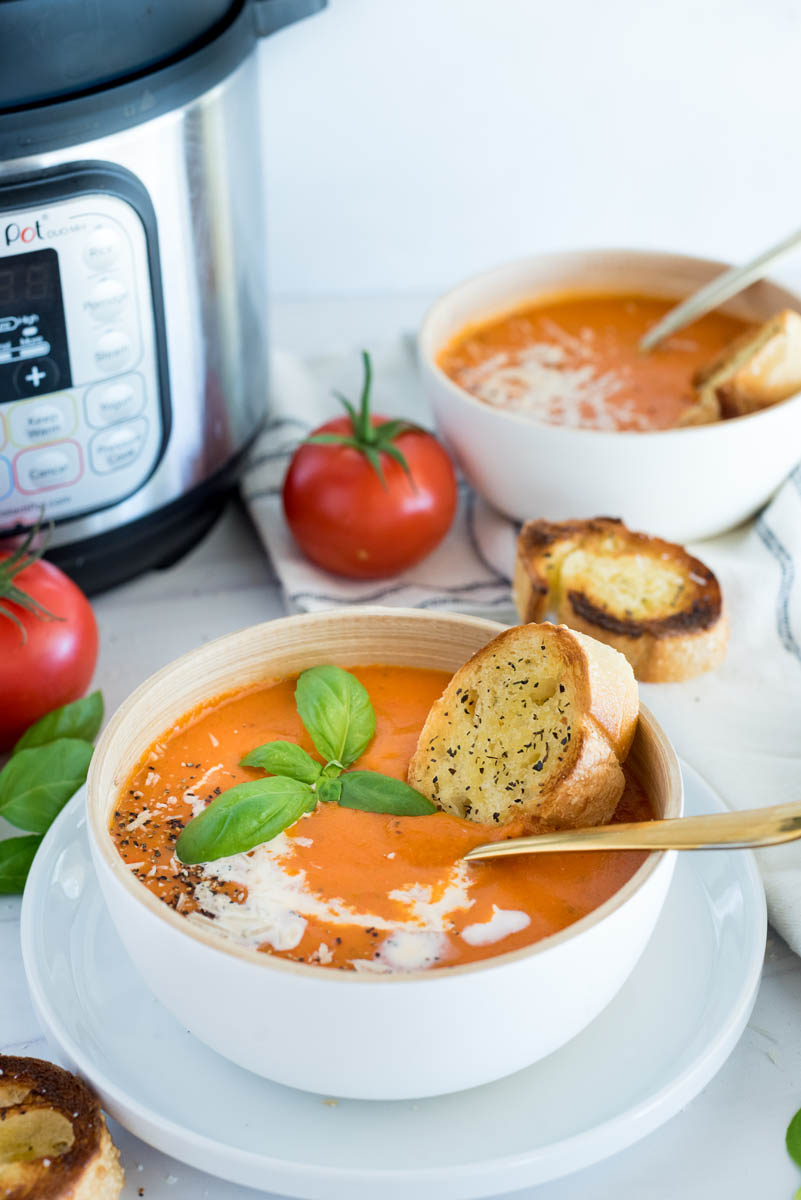 A 45 degree image of a white bowl filled with tomato basil soup, with an Instant Pot and fresh tomatoes in the background