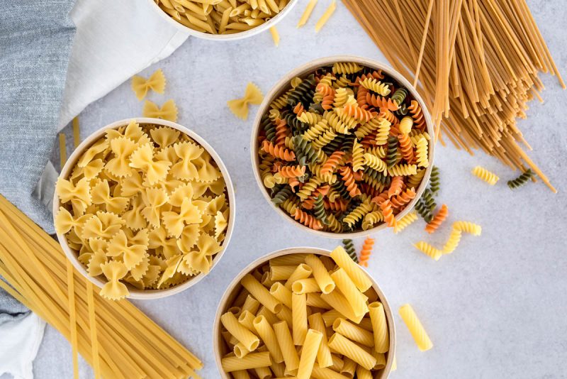 Overhead shot on three white bowls of pasta filled with bow ties, rotini, and penne pasta arranged in a triangular shape, with fettuccini  and spaghetti in the corners
