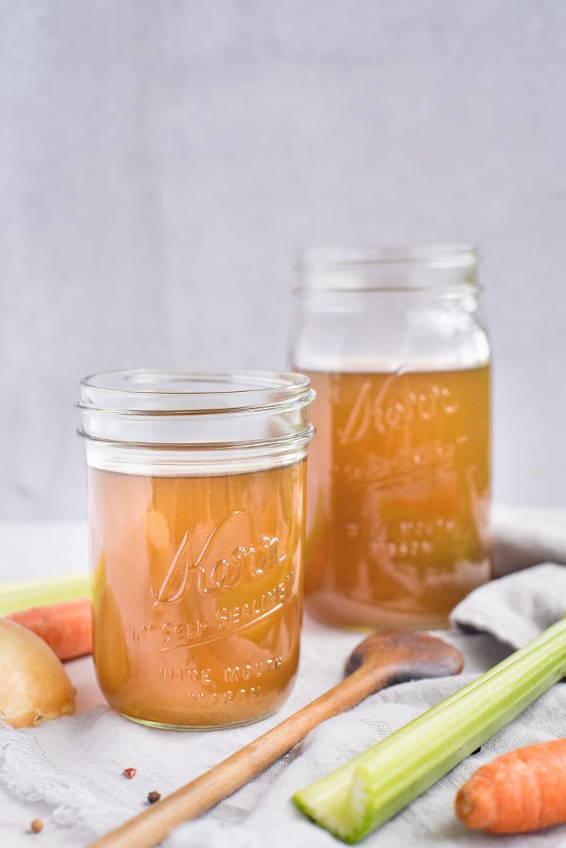 a vertical shot of two mason jars filled with fresh homemade vegetable broth made in an InstaPot pressure cooker, with a stick of celery and a carrot and wooden spoon in the foreground
