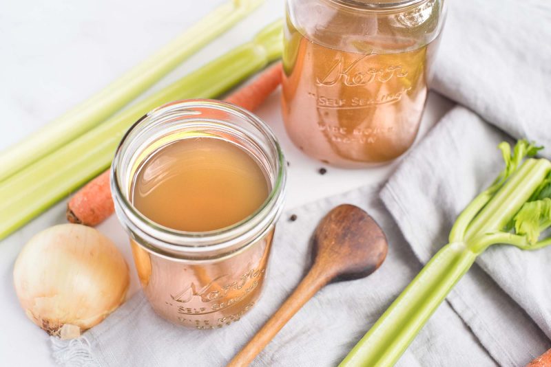 a 45 degree shot of two mason jars filled with vegetable broth, with an onion, carrot, celery, and wooden spoon visible in the background