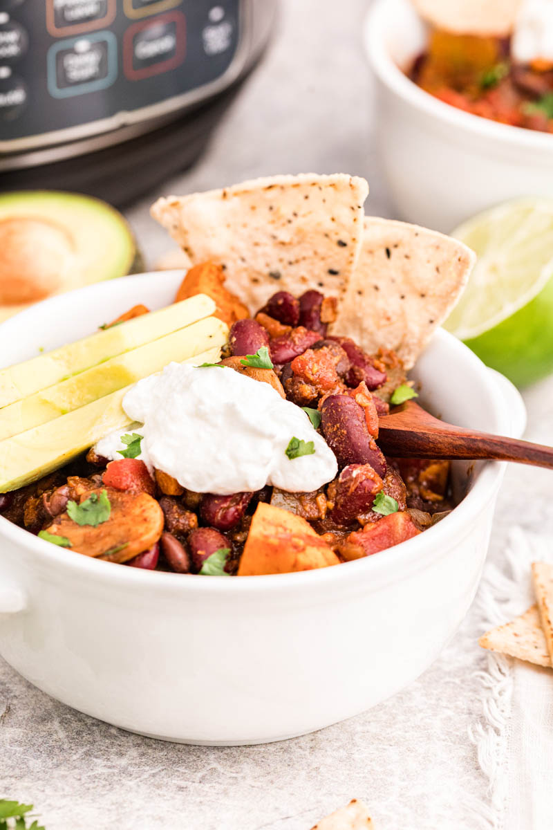 A vertical shot showing a white bowl full of vegetarian chili in front of an Instant Pot, garnished with sour cream, tortilla chips, and sliced avocados, with fresh cut lime and a small bowl of additional chili in the background.