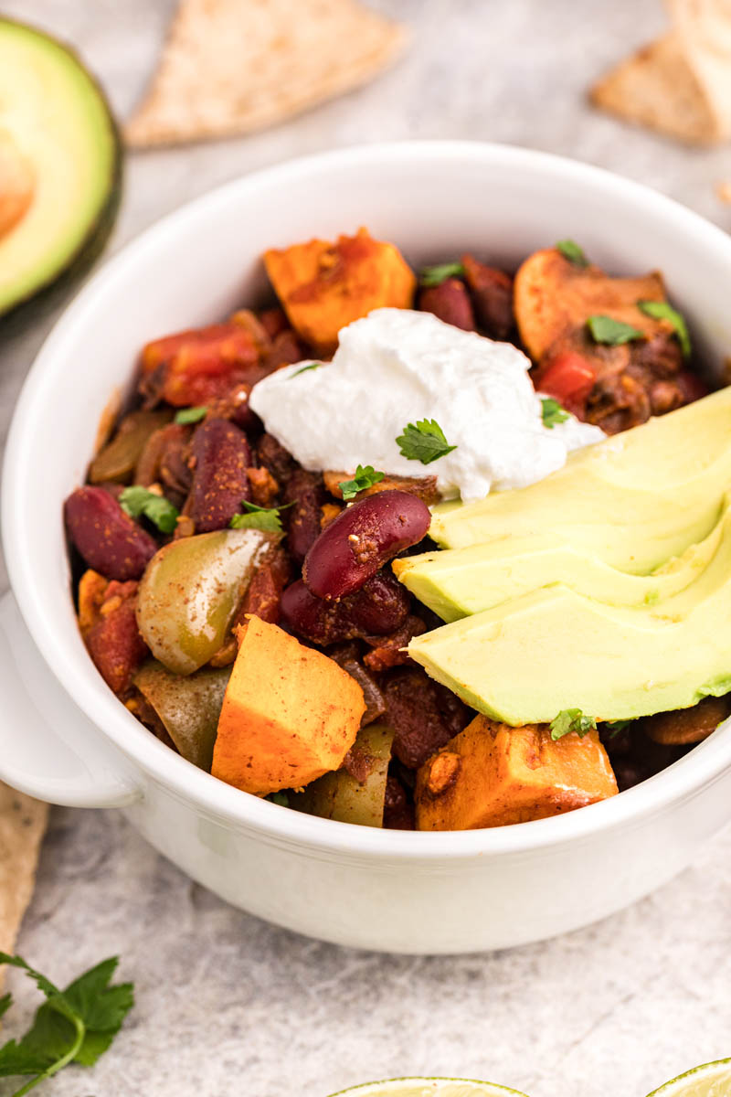 An overhead shot of the sweet potato chili  garnished with sour cream and avocados with a halved avocado and tortilla chips in the background