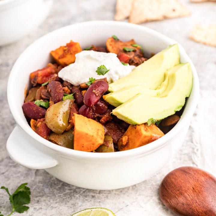 A 45 degree shot of Instant Pot Vegetarian Chili with sweet potatoes, mushrooms, and red kidney beans visible underneath a dollop of sour cream and sliced avocados