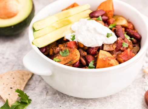 A 45 degree shot of vegetarian chili with sweet potatoes, beans, and mushrooms visible in the chili, topped with sliced avocados and sour cream and diced cilantro