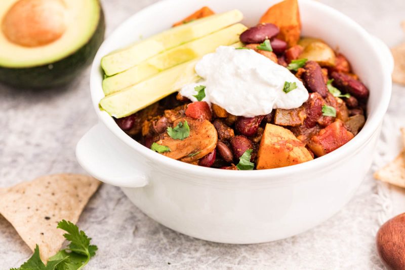 A 45 degree shot of vegetarian chili with sweet potatoes, beans, and mushrooms visible in the chili, topped with sliced avocados and sour cream and diced cilantro