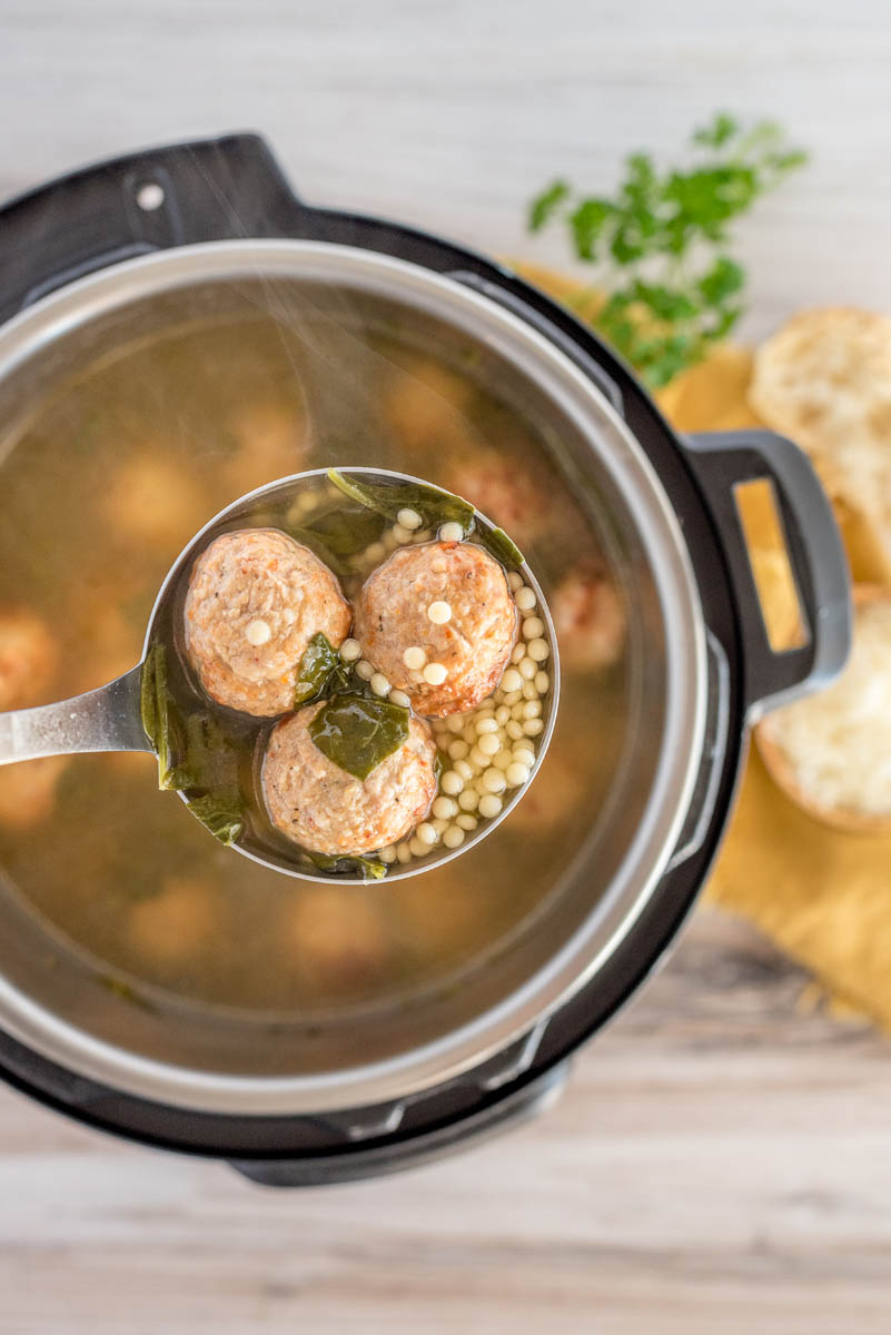 a vertical photo of a ladle full of Italian meatball soup with spinach and acini di pepe suspended over an Instant Pot filled with broth.