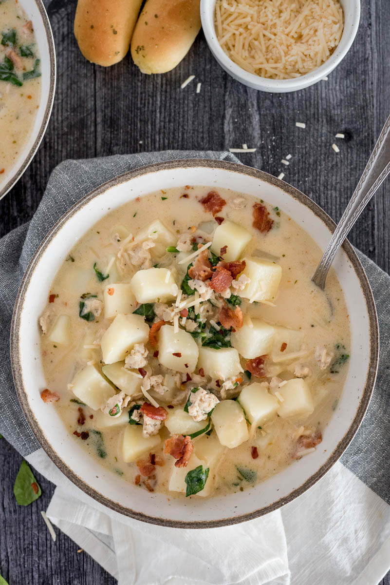 An overhead shot of a bowlful of zuppa toscana sitting on a green and white napkin with breadsticks and extra parmesan for garnish